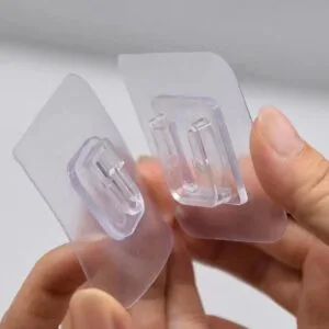 Gancho adhesivo transparente de doble cara sostenido con los dedos, listo para instalar
